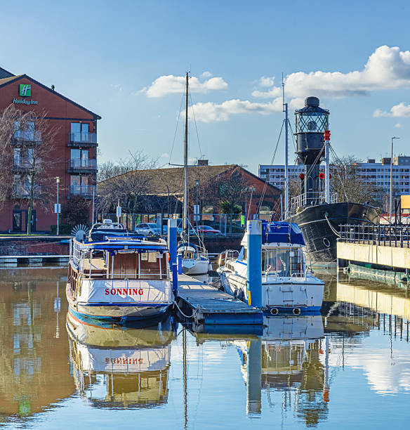 Hull, UK.  February 28, 2025.  Two boats are moored by a walkway and behind them is a light ship. There are buildings in the background and a sky with clouds above.