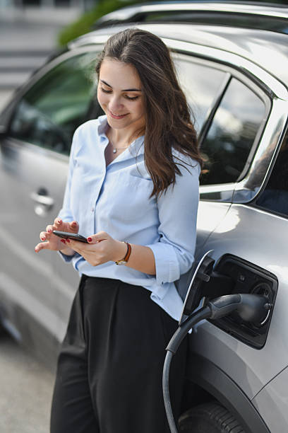 istock Happy Woman Using Smartphone While Charging Her Electric Vehicle Leaning on the Car 2207353443