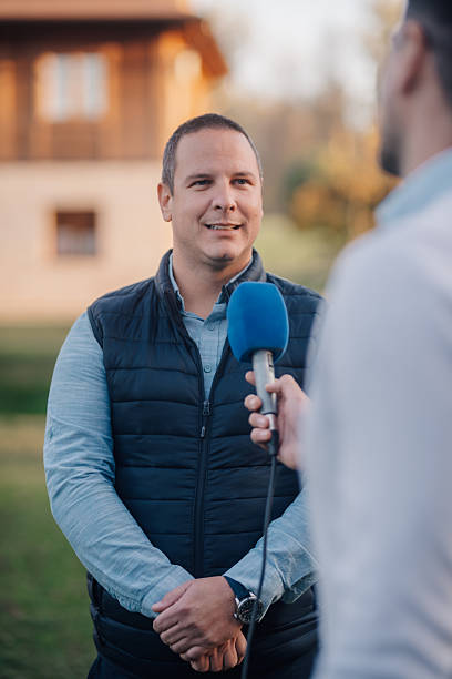 A male television reporter holding a microphone, is standing outdoors in front of a building. A male camera operator is filming him. Selective focus.