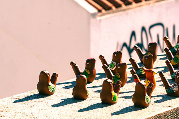 A collection of colorful handcrafted clay bird whistles arranged on a rooftop ledge in bright sunlight. Each whistle features hand-painted details, with one unique orange whistle standing out from the group. The setting is urban with graffiti in the background, adding contrast between folk art and street culture. Ideal for themes of individuality, artisan craft, and cultural creativity.