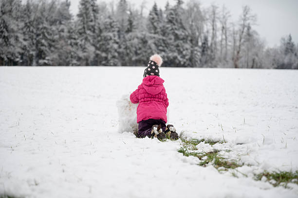 A child in a pink winter coat and polka-dot hat kneels in the snow, building a snowman in a peaceful, snowy landscape with a forest in the background.