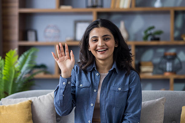 istock A smiling woman waves hello from her sofa in a bright, inviting, modern living room. 2207379677