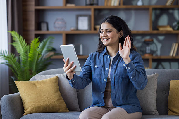 Smiling woman waving during a video call using her tablet while relaxing on the sofa at home.