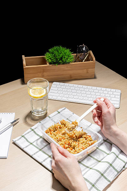 istock Office environment, break for nourishment. Snack at the workplace. Container of fried rice with carrots and chicken placed on a striped cloth. A hand holds a plastic fork. A glass of water with lemon. 2207384913