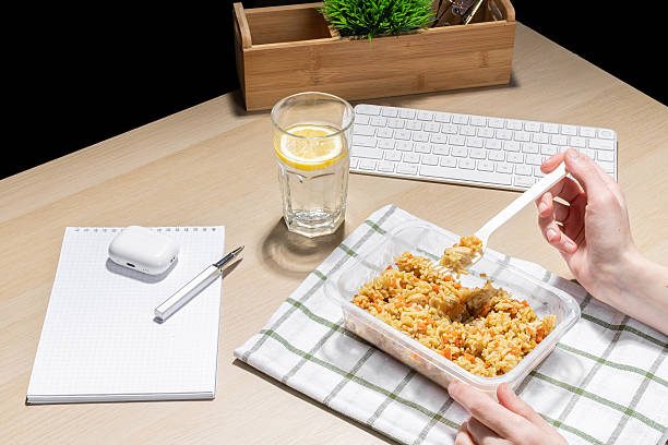 istock Office environment, break for nourishment. Snack at the workplace. Container of fried rice with carrots and chicken placed on a striped cloth. A hand holds a plastic fork. A glass of water with lemon. 2207384924