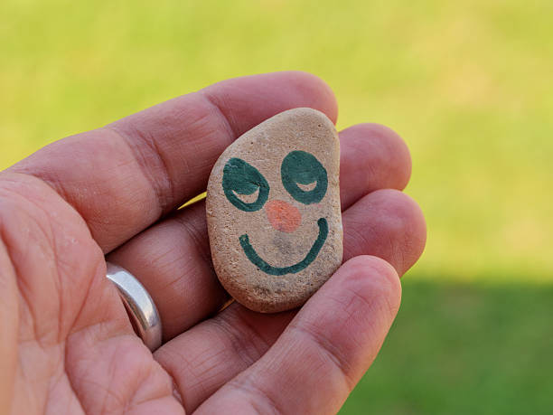 A hand grasps a stone adorned with a cheerful smiling face design.