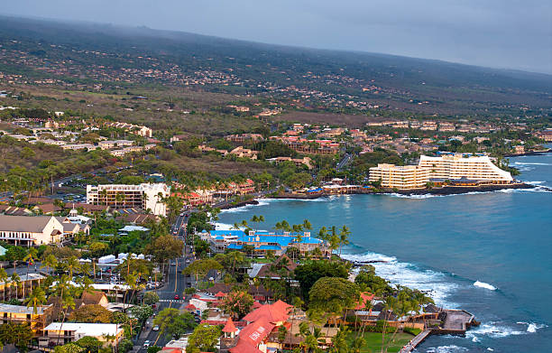 his drone photograph captures a stunning aerial view of a coastal town on the Big Island of Hawaii, featuring a double rainbow arching over the landscape. The image showcases the town's buildings, the coastline, and the vibrant colors of the rainbow against a dramatic sky.
