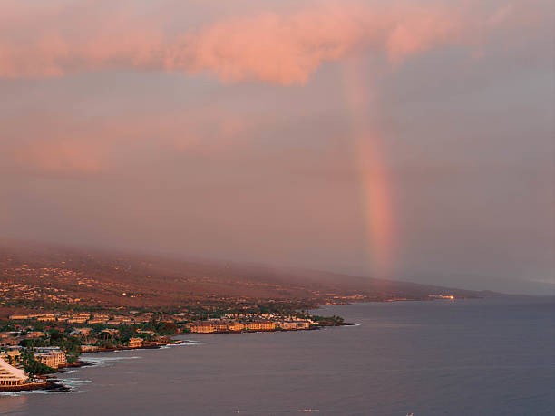 his drone photograph captures a stunning aerial view of a coastal town on the Big Island of Hawaii, featuring a double rainbow arching over the landscape. The image showcases the town's buildings, the coastline, and the vibrant colors of the rainbow against a dramatic sky.