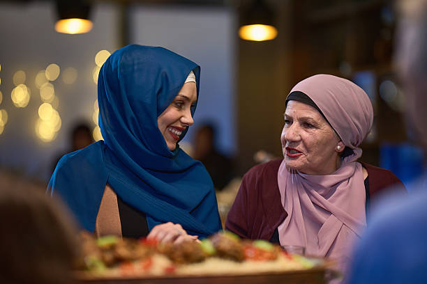 In a modern restaurant setting, a woman in hijab engages in conversation with her mother while they wait for the iftar meal, embodying familial bonding and cultural tradition during Ramadan.