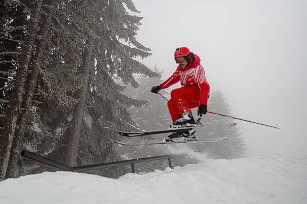 A skier in a vibrant red outfit performs a jump on a snowy mountain slope amidst mist-shrouded pine trees. The dynamic action emphasizes winter sports and adventure in a picturesque alpine setting, capturing wintertime excitement.