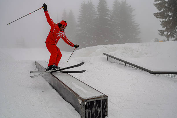 A skilled skier in bright gear performs a jump trick on a rail in a snowy setting. The scene depicts action and precision in winter sports as the skier expertly showcases their technique. Surrounding the action are snow-covered trees and fog, creating a serene and dramatic atmosphere.