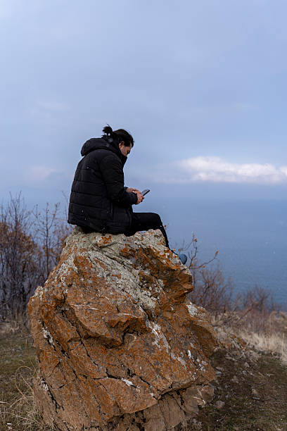 istock Man sitting on a rocky hilltop using a smartphone, winter landscape with ocean view, solitude and technology in nature, concept of digital nomad and remote work in cold weather 2207413573