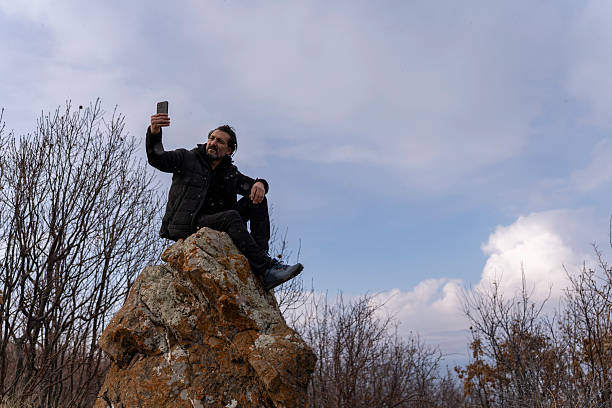 A man dressed in a black winter jacket sits on a rugged rock formation, engrossed in his smartphone. The backdrop features a vast ocean and a cloudy sky, with dry trees framing the scene. The winter atmosphere adds a sense of solitude and reflection, capturing the balance between technology and nature.