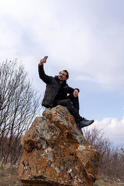 A man dressed in a black winter jacket sits on a rugged rock formation, engrossed in his smartphone. The backdrop features a vast ocean and a cloudy sky, with dry trees framing the scene. The winter atmosphere adds a sense of solitude and reflection, capturing the balance between technology and nature.