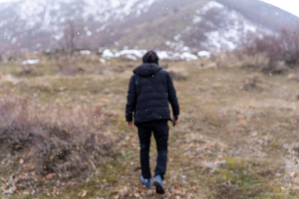 A man dressed in a black winter jacket walks through an open field towards a mountain with patches of snow. The sparse vegetation and dry grass create a raw and untouched wilderness atmosphere. Snowflakes gently fall, emphasizing the cold and remote environment, highlighting themes of exploration and resilience.