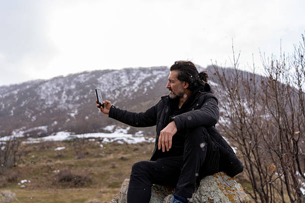 A man dressed in a black winter jacket sits on a rugged rock formation, engrossed in his smartphone. The backdrop features a vast ocean and a cloudy sky, with dry trees framing the scene. The winter atmosphere adds a sense of solitude and reflection, capturing the balance between technology and nature.