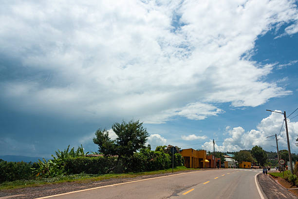 A quiet, paved road curves through the Rwandan countryside under a dramatic sky. On the left, lush green vegetation lines the roadside, while on the right, colorful buildings dot the landscape. A large, dark storm cloud looms in the distance, contrasting with patches of blue sky overhead. This image captures the serene beauty of rural Rwanda and the dynamic weather patterns of the region.