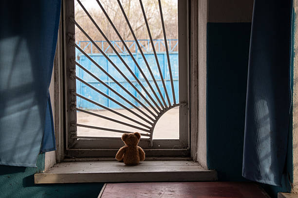 Children's brown bear sitting on a window with bars and looking out the window in an old abandoned house in Ukraine, abandoned