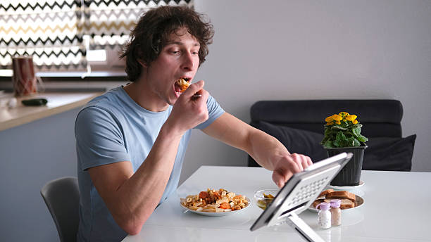 istock A cute guy is eating pasta with vegetables at a white table. While eating, he looks at something on the tablet. A delicious student's lunch 2207452839