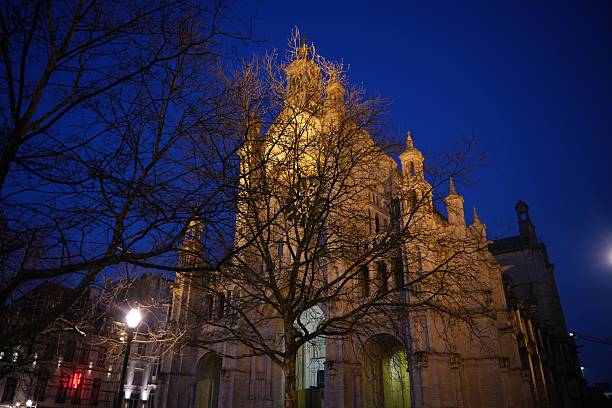 Evening view of Saint Catherine Church in Brussels, softly illuminated and partially hidden by leafless branches under a deep blue sky.