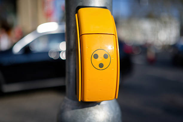 A yellow button with the three-dot symbol for blind and visually impaired people at a pedestrian crossing in an urban environment in a blurred background