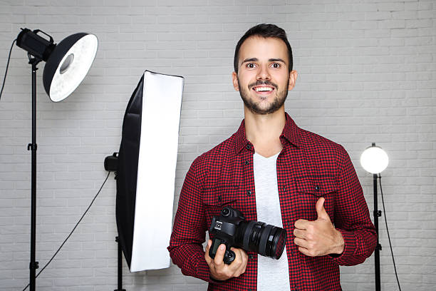 istock Young photographer showing thumb up with camera and professional studio equipment on grey background 2207467980