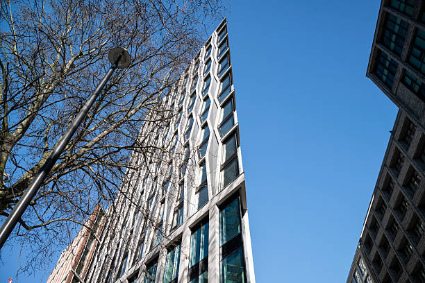 A sharply angled modern building in London with a striking geometric facade. The interplay of light, shadow, and reflections contrasts with the natural elements of a tree and the rigid lines of surrounding architecture.