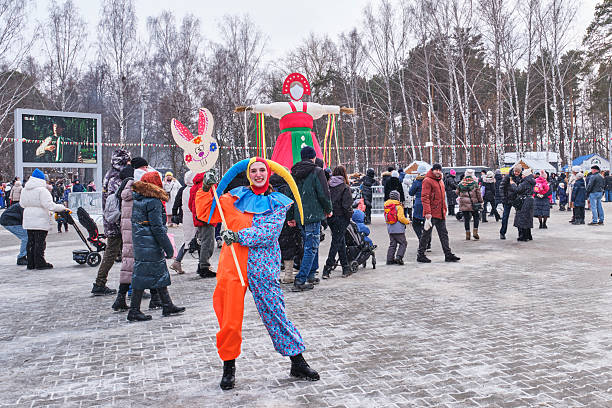 Yekaterinburg, Russia - March 2, 2025: People celebrating Maslenitsa in snowy park. Colorful clown performer, traditional effigy, and families enjoying festive winter atmosphere.