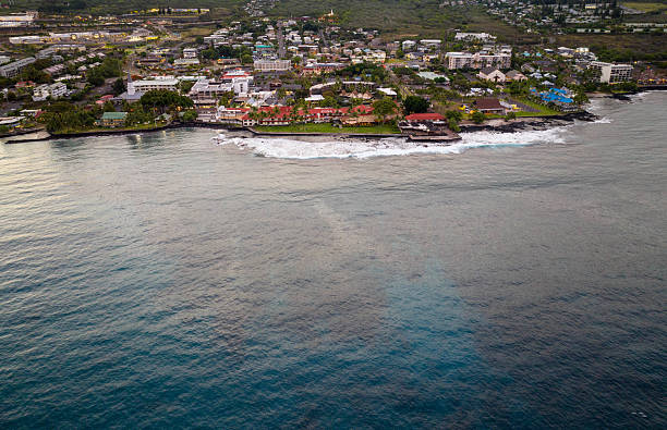 This drone shot captures the breathtaking beauty of the Kona coastline on Hawaii's Big Island, showcasing a stunning blend of urban life and the vast, tranquil Pacific Ocean. The warm hues of the setting sun cast a gentle glow over the scene, highlighting the coastal buildings nestled among lush greenery. The rhythmic waves crashing against the shore add a dynamic touch to this serene landscape.