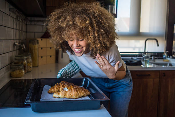 A young woman with natural curls stands in her minimalist kitchen, holding a tray of freshly baked croissants. With her eyes closed and a gentle smile, she takes in their irresistible aroma