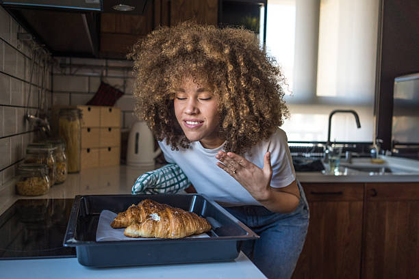 A young woman with natural curls stands in her minimalist kitchen, holding a tray of freshly baked croissants. With her eyes closed and a gentle smile, she takes in their irresistible aroma