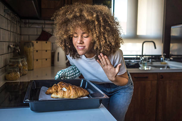 A young woman with natural curls stands in her minimalist kitchen, holding a tray of freshly baked croissants. With her eyes closed and a gentle smile, she takes in their irresistible aroma