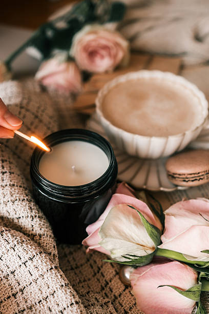 istock A cozy scene with a hand lighting a candle using a matchstick, surrounded by soft beige fabric, a cup of frothy coffee, pink roses, and a macaron. Warm and inviting atmosphere 2207512338
