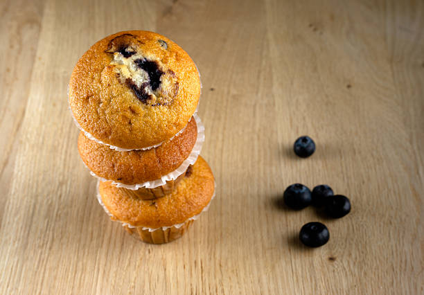 Five freshly baked blueberry muffins on a blue countertop, surrounded by blueberries and flour scattered around.