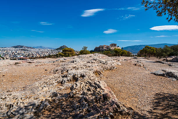 Panoramic view of the Acropolis and Athens city from a rocky hill, showing ancient ruins and modern buildings under a blue sky.