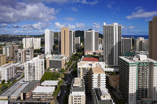 A stunning aerial view of the Waikiki skyline in Honolulu, Hawaii, with the iconic Diamond Head crater rising in the background. The image showcases the dense cluster of modern high-rise hotels, apartments, and commercial buildings under a bright blue sky. The Pacific Ocean peeks through the horizon, adding a tropical touch to the vibrant urban landscape. A perfect snapshot of the unique blend of city life and natural beauty in Oahu.