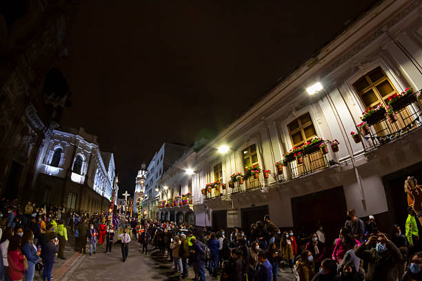 Quito, Pichincha, Ecuador - April 14, 2022: Crowd of people wearing face masks watching a procession of hooded penitents carrying a statue of Jesus Christ during Holy Week in Quito, Ecuador