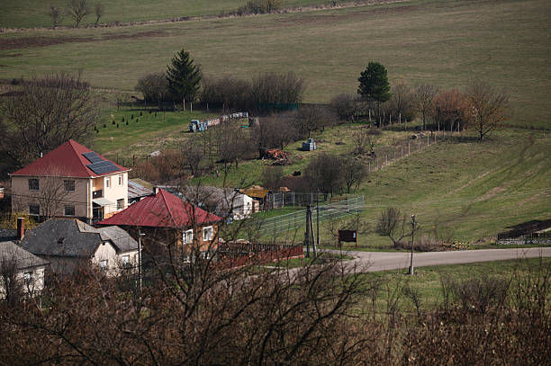 A cluster of detached houses sits nestled in a quiet valley, surrounded by grassy hills and bare trees. The early spring setting and soft natural light create a peaceful, pastoral atmosphere typical of countryside living.