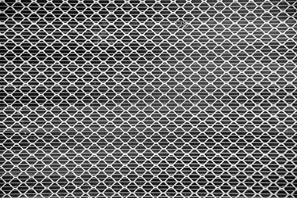 A detailed black and white image showing a repetitive diamond-shaped metal mesh pattern. The strong geometric lines and monochrome tones create a striking industrial texture and abstract visual rhythm.
