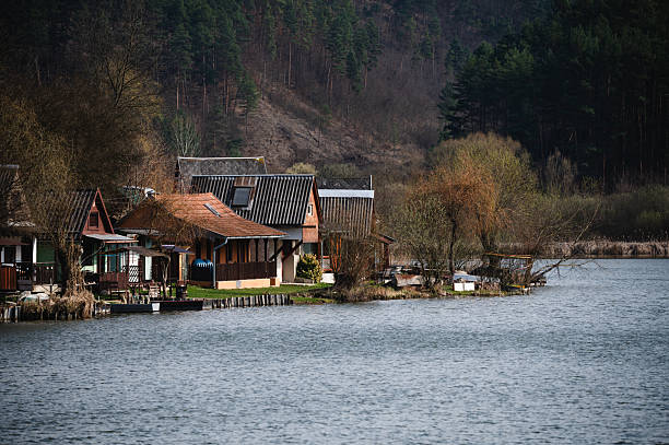 Charming wooden cottages line the edge of a calm lake, backed by forested hills and bare early-spring trees. The peaceful lakeside scene reflects a quiet, rustic atmosphere ideal for retreat and relaxation in nature.