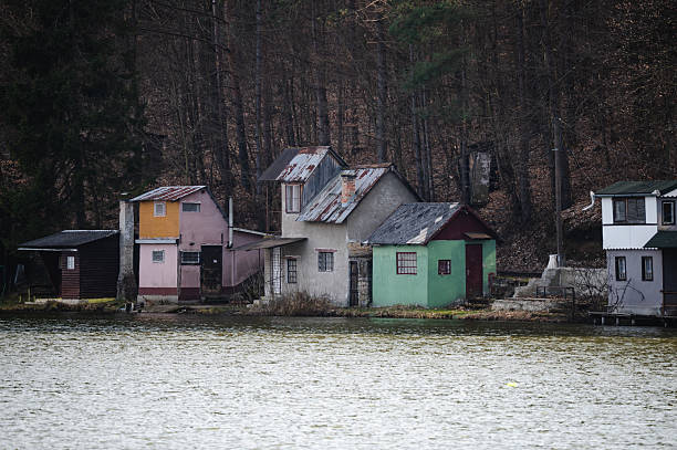 Charming wooden cottages line the edge of a calm lake, backed by forested hills and bare early-spring trees. The peaceful lakeside scene reflects a quiet, rustic atmosphere ideal for retreat and relaxation in nature.