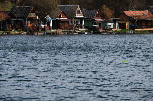 Charming wooden cottages line the edge of a calm lake, backed by forested hills and bare early-spring trees. The peaceful lakeside scene reflects a quiet, rustic atmosphere ideal for retreat and relaxation in nature.