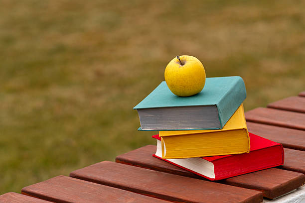 Tilted stack of books with yellow apple placed outdoors.