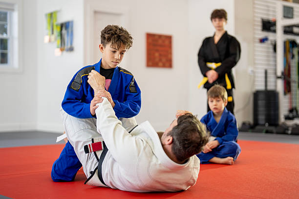 Focused adult man learning effective self defence techniques in training sparring, practicing painful armlock on male opponent lying face down on floor in gym