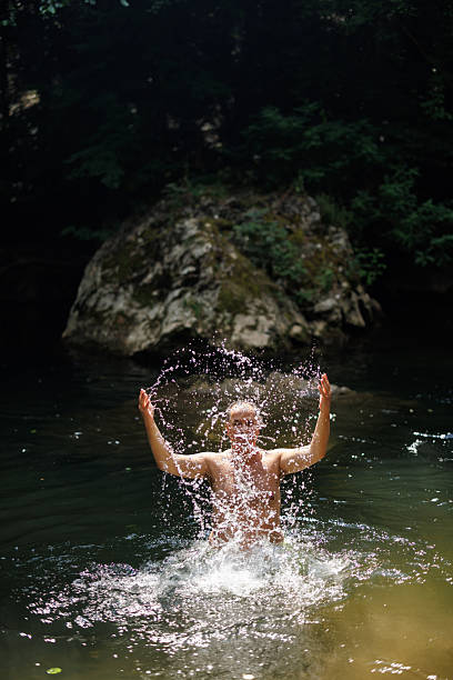 Playful mature man standing in the shallow of small river and splashing water on sunny summer day