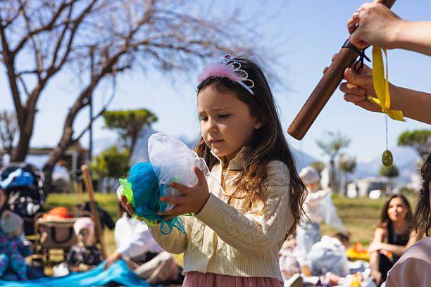 A young girl wearing a tiara examines a colorful craft object in a sunny park. Other children and families are visible in the background, enjoying various outdoor activities during the day picnic.