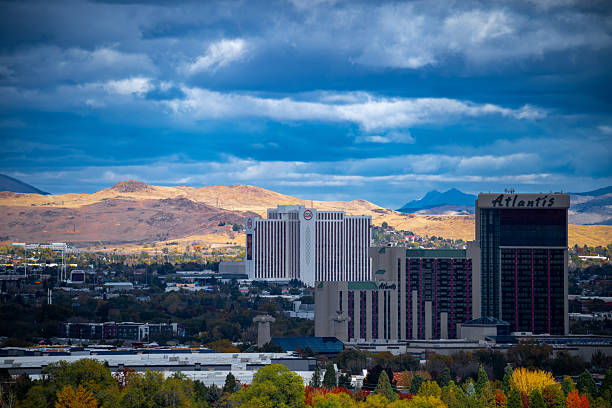 A dramatic view of the Atlantis Casino Resort Spa in Reno, Nevada, with golden high desert hills and a patchwork of colorful autumn trees in the foreground. The cloudy sky casts a moody light over the scene, enhancing the contrast between the illuminated buildings and the surrounding landscape.