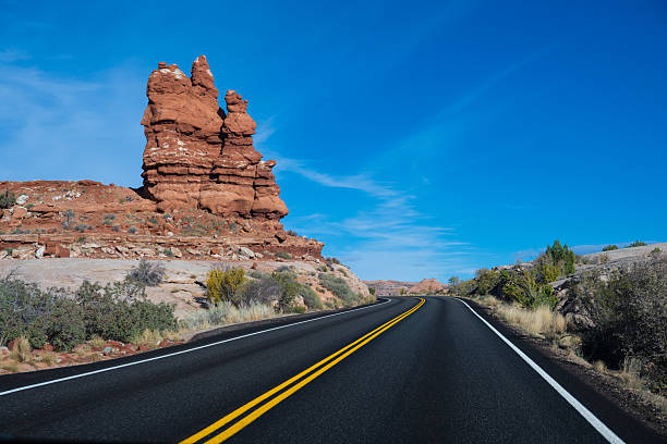 A winding highway cuts through the stunning red rock formations of Arches National Park, creating a perfect scene for road trips and adventure travel. The smooth asphalt road, flanked by desert vegetation and sandstone cliffs, leads toward the iconic rock landscapes of Utah, under a clear blue sky. This image captures the freedom of the open road, making it ideal for themes of exploration, travel, and scenic drives through the American Southwest.