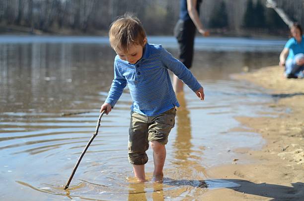 young child playing with stick by water's edge - lakeside sosyal tesisleri stok fotoğraflar ve resimler
