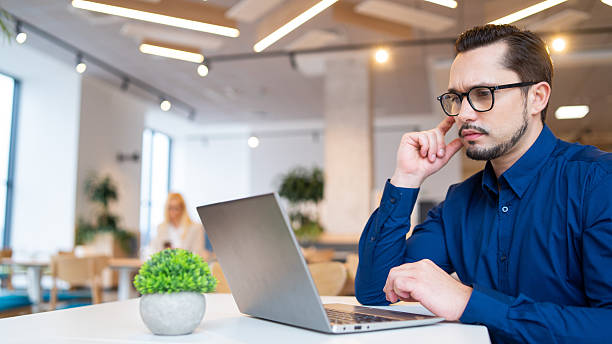 istock Man with thoughtful expression using laptop in lively workspace, enhancing productivity and focus. 2207623015